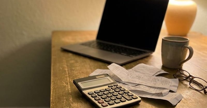 A pocket calculator and a scatter of paper receipts beside a laptop under a warm lamp